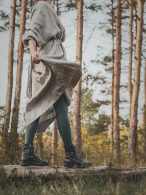 Uzun gri elbiseli, hareket bulanıklığı ve yeşil taytlı bir kadının, sonbaharda bir çam ağacının ormanında ağaç kütüğünde yürüyüşünün yan görüntüsü.