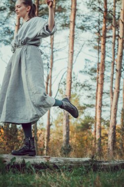 Beyaz tenli, uzun organik keten elbiseli bir kadının portresi bir çam ormanındaki ağaç kütüğünün üzerinde dengede duruyor.: yavaş moda, minimalizm, el yapımı giysiler, doğal güzellik.