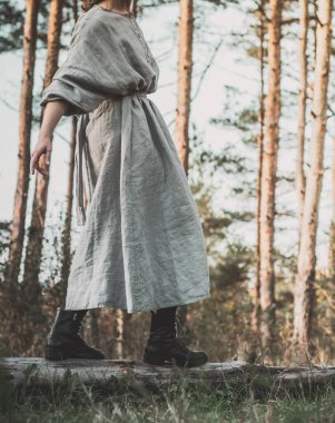 Arka planda çam ağacı ormanı olan uzun organik keten elbiseli beyaz kadın portresi. Yavaş moda, minimalizm, el yapımı kıyafetler, doğal güzellik.