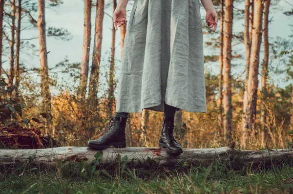 Lower body front view of woman in a long grey linen dress and black leather boots on a fallen pine tree log with coniferous tress in the background
