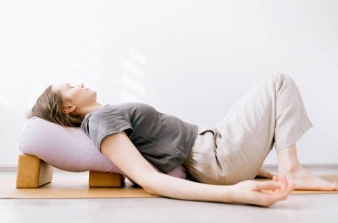 Günlük kıyafet giymiş bir kadın bloklarla desteklenen yin / restoratif yoga yapıyor.