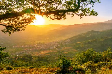 Dağlarda harika bir bahar günbatımı. Yeşil ağaç dalları arasında dağlardan harika bir manzara. Kasaba, dağlar ve arka planda güzel bir günbatımı olan bir vadiye.