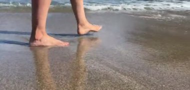 Kumsalda yalın ayak yürüyen bir kadın, dalgalar ayaklarının üzerinde yankılanıyor.