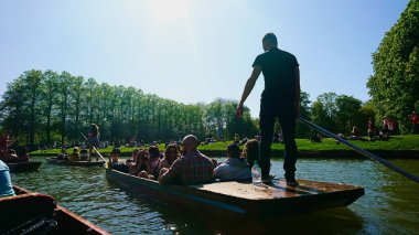 Cambridge, Birleşik Krallık - 5 Mayıs 2018: Güneşli bir günde, öğrenciler salgından önce çayırda ve ormanda piknik yaparken, Nehir Kamerası 'nda sörf yapan genç bir adamın sırtı