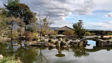 Kagoshima, Japonya - 01.29.2020: Sengan-en 'de bir gölet üzerindeki taş köprü ve ağaçların yansıması ve Sakurajima' nın arkadaki mavi gökyüzünün altında duman yayması