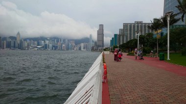 Hong Kong - 08.01.2021: Kowloon 'da Wai Chai ve Kowloon' da Wai Chai ile Kowloon Limanı boyunca Victoria Limanı boyunca yürüyen yolcular bulutlu bir gökyüzünün altında.