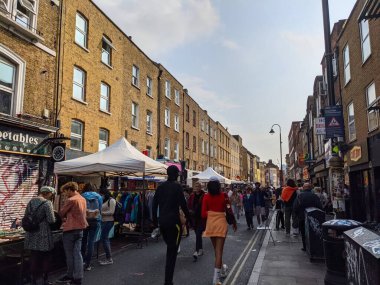 Londra, İngiltere - 09.26.2021: Kalabalık Brick Lane açık hava pazarında gezen ziyaretçiler, güneşli bir günde mavi gökyüzü altında satıcılar ve tezgahlarla yan yana duruyorlar.