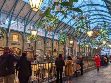 Londra, İngiltere - 11.05.2021: Parmaklıkların yanında duran ziyaretçiler, alacakaranlıkta eğimli cam bir tavan altında Greko-Romen tarzı Covent Garden pazarında bir restoran oturma alanına bakıyorlar.