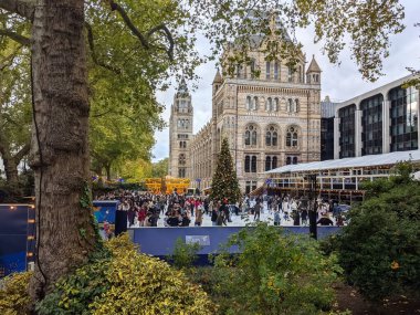 Londra, İngiltere - 11.06.2021: İnsanlar Doğal Tarih Müzesi Buz Pisti 'nde kafenin ve barın yanında bir Noel ağacı ve bulutlu bir gökyüzünün altında arkada atlıkarınca ile buz pateni yapıyorlar.