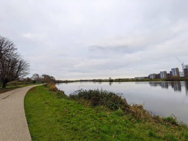 London, UK - 12.16.2021: An empty pathway next to a lake in the Walthamstow Wetlands along trees and marshes under a cloudy sky with buildings at the edge clipart