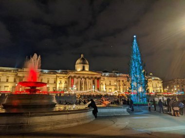 Londra, İngiltere - 12.21.2021: Trafalgar Meydanı 'ndaki işlek bir açık hava Noel pazarı, Norveç tarafından gönderilen aydınlatılmış Noel ağacı ve gece vakti bir çeşme ile Ulusal Galeri önünde.