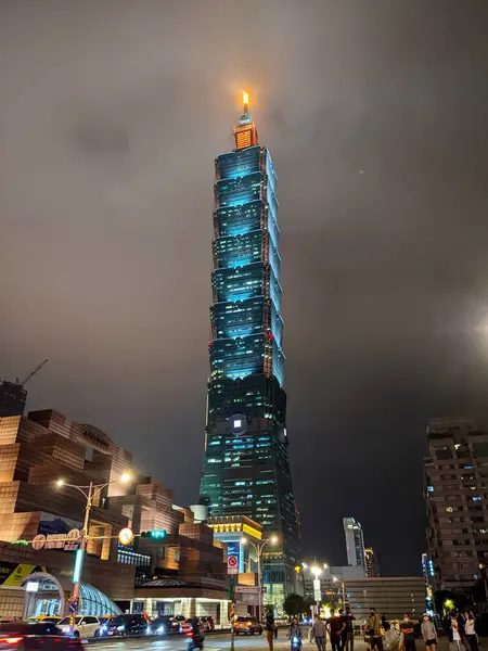 stock image Taipei, Taiwan - 11.22.2022: The illuminated Tapei 101 with the Taipei World Trade Center and a MRT exit in the front and cars and pedestrians on Xinyi Road at night during the pandemic among mist