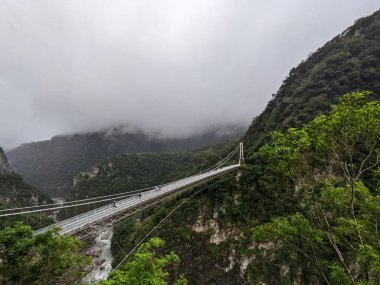 Taroko, Tayvan - 11.26.2022: Liwu Nehri boğazı üzerindeki Buluowan Kablo Askı Köprüsü üzerinde yürüyen turistlerin yağmurlu bir günde salgın sırasında meydana gelen 403 depreminden önce dağlardaki sisli manzarası
