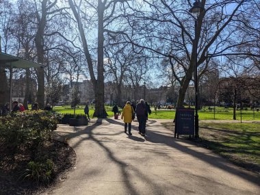 Londra, İngiltere - 03.18.2022: İnsanlar salgın sırasında açık mavi bir gökyüzünün altındaki banklarda oturan diğerleriyle birlikte çayırlardan geçen bir kafenin yanındaki kafeye yürüyorlar