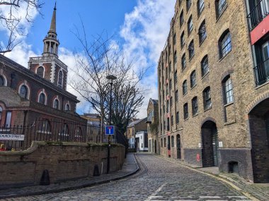 Londra, İngiltere - 2023.03.22: St. Marys Kilisesi, Rotherhithe 'nin yanında kaldırım taşı olan ve güneşli bir günde mavi gökyüzü altında tuğla fabrikalarını dönüştüren boş bir cadde