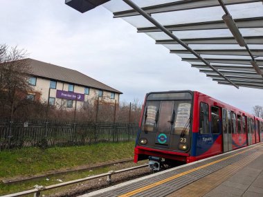 Londra, İngiltere - 2023.03.11: Kırmızı ve mavi bir DLR treni Beckton İstasyonu 'na boş bir platformla yaklaşıyor arkasında bir Premier Inn, cam bir gölgelik ve bulutlu bir gökyüzü var.