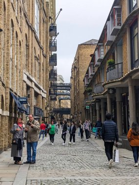 Londra, İngiltere - 2023.05.13: Tower Bridge 'in yanındaki tarihi nehir kenarı sokağı olan Shad Thames' te yürüyen yayalar, dönüştürülmüş depo kompleksi, köprüler altındaki dükkanlar ve bulutlu bir gökyüzü