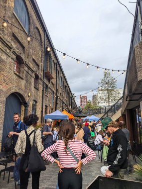 Londra, İngiltere - 2023.10.01: Coal Drops Yard 'daki işlek bir sokakta, pazar tezgahları ve dükkanların yanında, bulutlu bir gökyüzünün altında yiyecek ve içecek satan insanlarla birlikte yürüyen ziyaretçiler