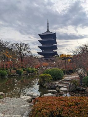 Kyoto, Japonya - 2023.12.14: Toji beş katlı huzurlu bir sonbahar bahçesinin üzerinde yansıtıcı bir gölet ve Toji 'de iyi manikürlü çalılar bulutlu bir gökyüzünün altında güneş ışığı bulutları üzerinde siluet oluşturur.