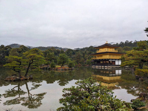 Киото, Япония - 2023.12.15: Золотой павильон в Кинкаку-дзи с культивируемыми соснами спереди, отражением на пруду и лесом сзади под облачным небом