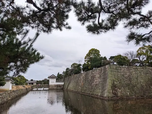 Kyoto, Japonya - 2023.12.16: Nijo Kalesi 'ndeki geniş hendeğin yanındaki iç taş duvarlar hendeği geçerek hendeği geçen bir köprü ile bulutlu bir gökyüzünün altındaki bahçedeki iyi korunmuş ağaçlar.