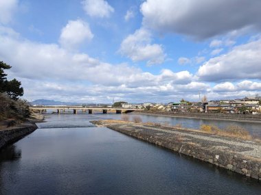 Uji, Japan - 12.17.2023: View of Uji Bridge across Uji River alongside riverside footpaths and trees with Uji Park in the middle of the river in an autumn afternoon under a blue sky with clouds clipart