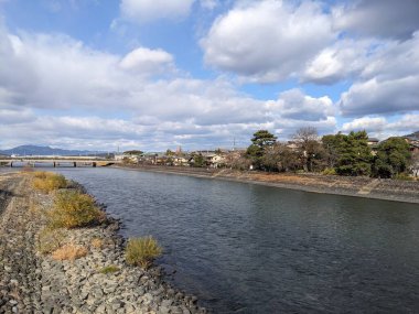 Uji, Japonya - 12.17.2023: Uji Nehri 'nin karşısındaki Uji Köprüsü' nün nehir kenarındaki patika ve ağaçlarla birlikte Uji Parkı 'nın ortasındaki mavi gökyüzü altında bulutların altında görüntüsü