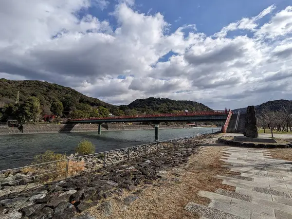 Uji, Japonya - 12.17.2023: Uji Nehri 'ni geçmek için Asagiri Köprüsü üzerinde yürüyen insanlar, diğer tarafta kırmızı bir Torii kapısı, Uji Parkı' ndaki patikadan görülebilen bulutlu mavi bir gökyüzü.