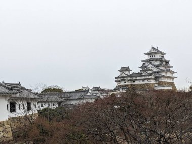 Himeji, Japonya - 12.19.2023: Himeji Kalesi ve çevresindeki yaguralar, veya taretler, bulutlu bir gökyüzünün altındaki bahçede sonbahar ağaçları ile