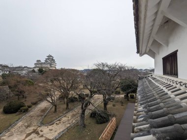 Himeji, Japonya - 12.19.2023: Himeji Kalesi 'nde, bahçesinde sonbahar ağaçları ve bulutlu bir gökyüzünün altında ana kale bulunan yagura ya da taretlerin çatılarına yakın çekim