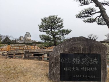 Himeji, Japonya - 12.19.2023: Kale kompleksine giden ahşap bir köprüyle Himeji Kalesi 'ne giriş, arkada Ana Kale ve bulutlu bir gökyüzünün altında isimlendirilmiş bir taş levha.