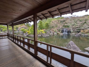 Himeji, Japan - 12.19.2023: A wooden observation deck under a traditional roof towards a peaceful pond with goldfishes surrounded by a waterfall and autumn trees in Koko-en without tourists clipart