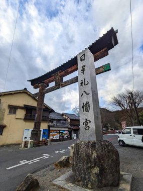 Omihachiman, Japan - 12.20.2023: A wooden torii gate and a stone stele engraved with the shrines name at the entrance of Himure Hachiman-gu with a road passing through under a cloudy blue sky clipart