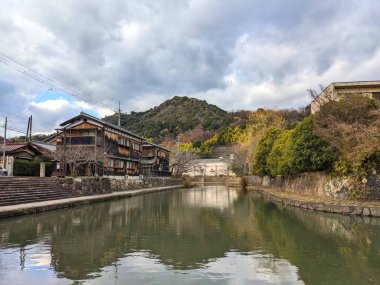 Omihachiman, Japonya - 12.20.2023: Geleneksel ahşap binalar ve sonbahar ağaçları Hachimanbori kanalı yanında bir dağın önünde turistler olmadan bulutlu mavi bir gökyüzünün altında