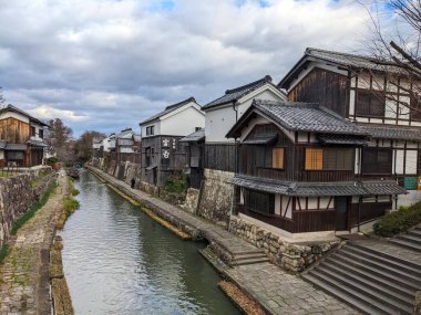 Omihachiman, Japonya - 12.20.2023: Hachimanbori su yolu kenarındaki tarihi binalar ve patikalar, turistler olmadan bulutlu bir gökyüzünün altında demirlemiş geleneksel tekneler