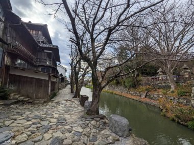 Omihachiman, Japonya - 12.20.2023: Hachimanbori su yolu boyunca, yapraksız ağaçların ve turistler olmadan bulutlu mavi gökyüzünün altında tarihi binaların yanında taşla döşenmiş bir patika
