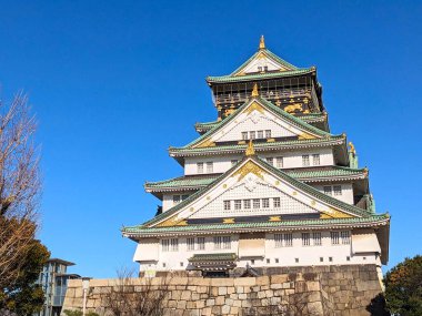 Osaka, Japonya - 12.21.2023: Osaka Kalesi 'nin cephesi altın süslemeleri, beyaz duvarı ve dulavratlı duvarın tepesindeki geleneksel çatısı açık mavi gökyüzü altında.