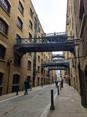 Londra, İngiltere - 05.13.2024: Shad Thames caddesinde yürüyen ve konuşan insanlar dönüştürülmüş depolar ve fayanslı zemin ve zemin kattaki mağazalar üzerindeki yükseltilmiş yürüyüş yolları bulutlu bir gökyüzü altında