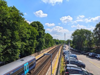 Meopham, UK - 06.05.2024: A Southeastern Railway train stopped at the platform of Meopham Station alongside trees and next to a full car park under a sunny blue sky clipart