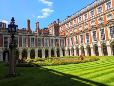 Londra, İngiltere - 07.19.2024: Hampton Court Palace 'daki Çeşme Mahkemesi kırmızı tuğlalı binalar ve mermer sütun koridorları ile şehir merkezinde güneş ışığı altında bir çeşme ile çevrili