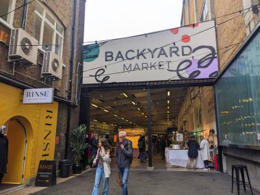 London, UK - 26.10.2024: Entrance to the indoor Backyard Market, which sells handicrafts with two customers just left the market under a cloudy sky next to brick-built buildings and a shop clipart