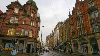 Manchester, İngiltere - 1.19.2018: Her iki tarafında dönüştürülmüş kırmızı tuğla binaları ve dükkanları olan sıradan bir cadde, kaldırımda yürüyen yayalar ve yaya geçitleri, bulutların altındaki tramvay rayları
