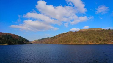 Peak District, İngiltere - 2.25.2018: Ladybower Reservoir 'ın diğer tarafındaki ormanlık tepelerin altında güneşli mavi gökyüzünün altında, sudaki bulutlarla huzurlu ve sakin bir manzarası