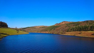 Peak District, İngiltere - 2.25.2018: Kışın turistler olmadan açık mavi gökyüzü altında ormanlı tepelerle çevrili sakin ve sakin mavi Ladybower Reservoir manzarası