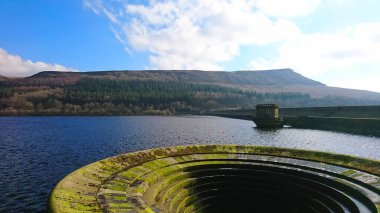 Peak District, UK - 2.25.2018: A dry and mossed spillway in the Ladybower Reservoir and the Ladybower Dam on the side with a hill at the back in the winter under a sunny blue sky without tourists clipart