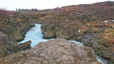 Iceland - 3.25.2018: Tourists watching Barnafoss on the River Hvita flow through rocky terrain and bare trees from an observation dock on the surrounding trail under a cloudy sky before the pandemic clipart