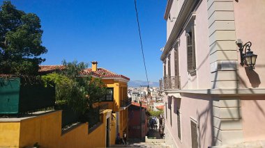 Atina, Yunanistan - 29,3,2018: Yamaçta, kentsel ufuk çizgisine ve ufuktaki dağlara doğru uzanan yerleşim yerleri boyunca merdivenlerde ve dükkanlarda oturan insanların oturduğu sıradan bir ara sokak