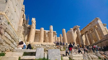 Athens, Greece - 29.3.2018: Tourists walking through the Propylaea, entrance to the Acropolis, among marble columns and other ruins under a clear blue sky with sunlight in summer before the pandemic clipart