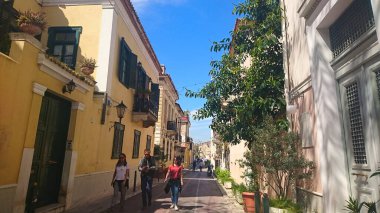 Atina, Yunanistan - 29,3,2018: Salgından önceki yaz güneşli açık mavi bir gökyüzünün altında, sakin bir sokakta renkli konutlarla yürüyen yayalar