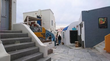 Santorini, Greece - 3.31.2018: Tourists walking on the narrow alley in Oia, browsing souvenir shops and handicrafts among colourful buildings under a cloudy sky before the pandemic clipart